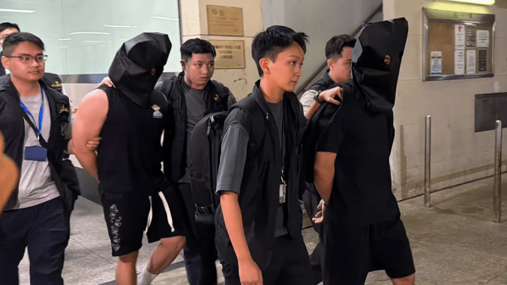 執法人員在觀塘工廠大廈單位外，將兩名被黑布蒙頭的被捕男子押解離開現場。