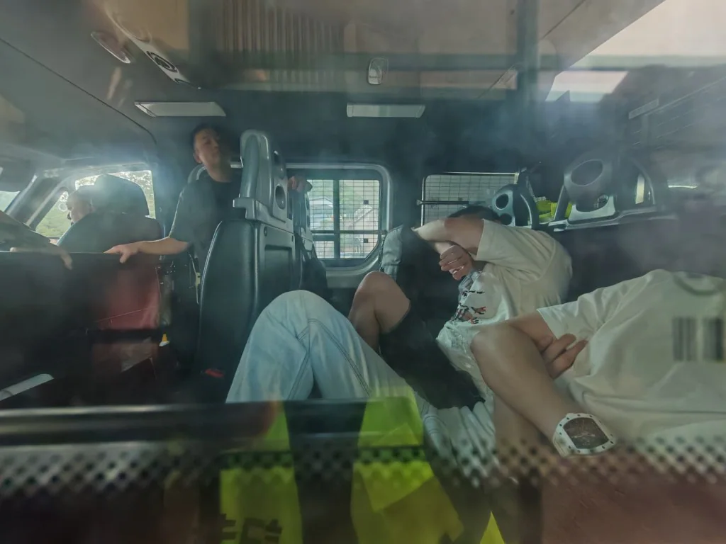 五名男子坐在警車車廂內，部分人以手掩面，正接受警方調查。