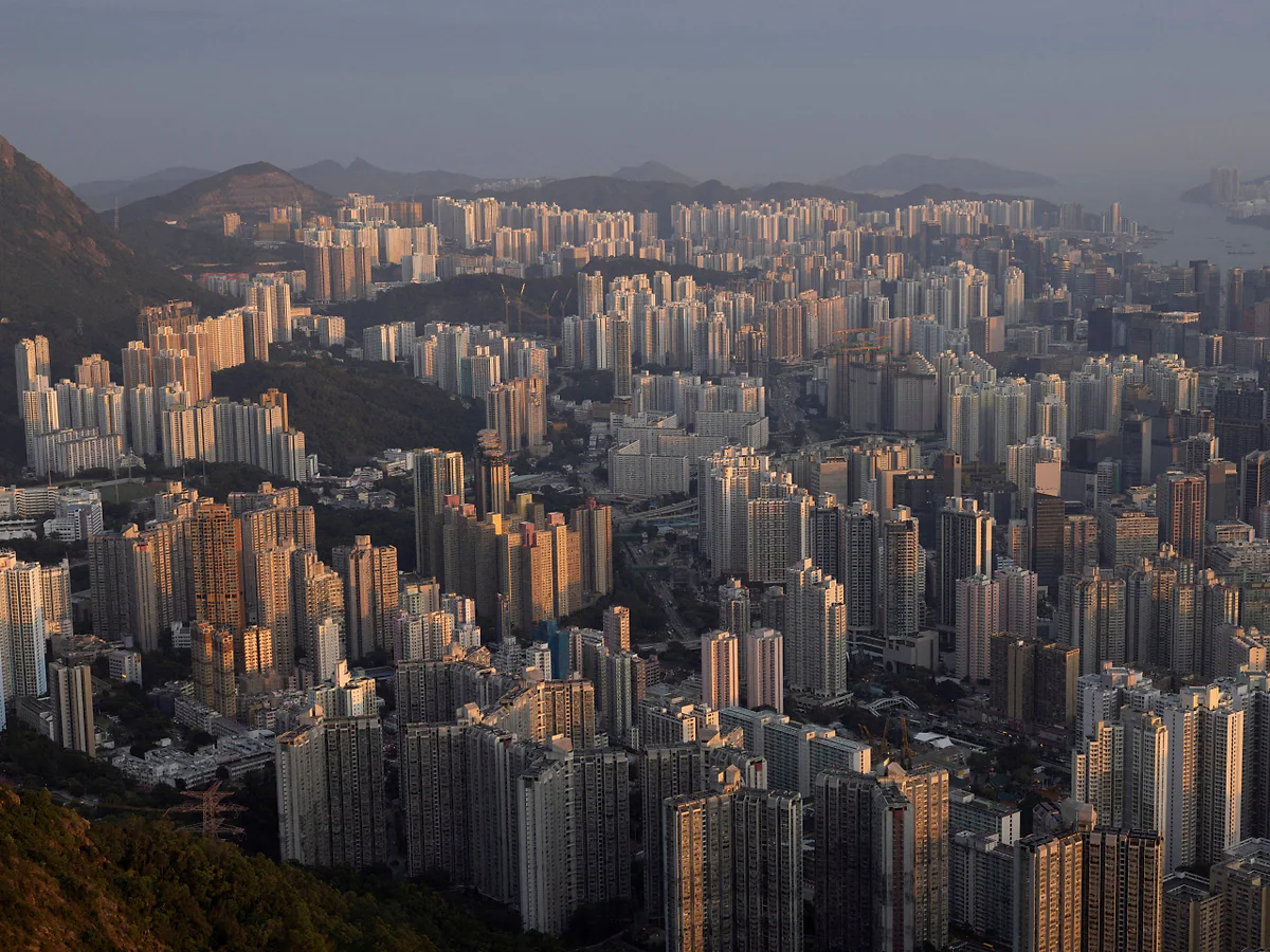 香港密集的住宅大廈群，建築物在陽光照射下呈現出深淺不一的色調，背景可見山巒及部分海港景觀。