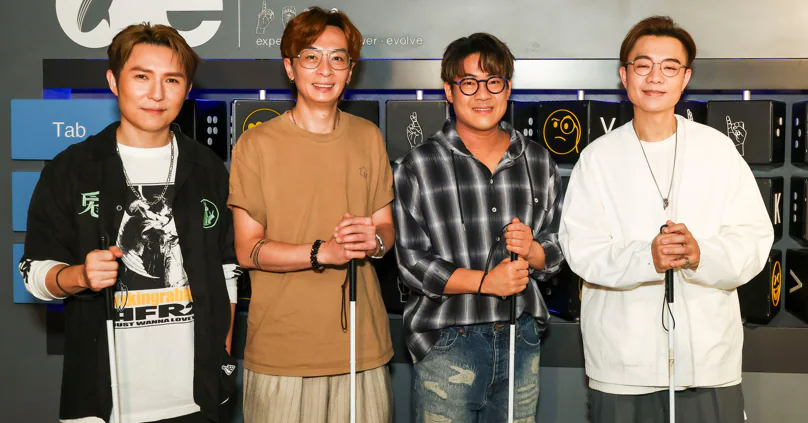 羅啟豪、劉威煌、顏志恒及梁浩賢於黑暗中對話體驗館合照，其中兩人手持白色導盲杖，背景為印有鍵盤符號的牆壁。