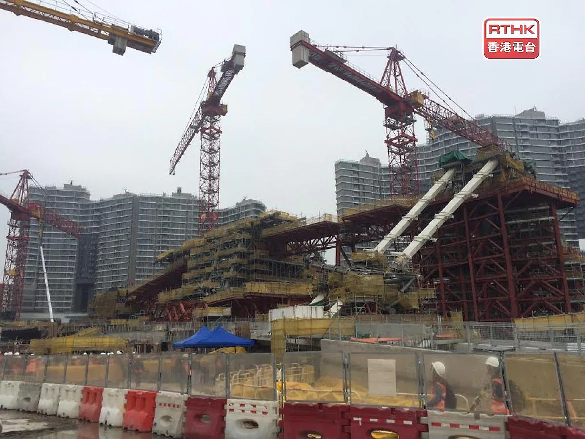 大型建築地盤設有多座塔式起重機及金屬支架，背景為住宅大廈，前方有身穿反光衣及佩戴安全帽的人員走過。