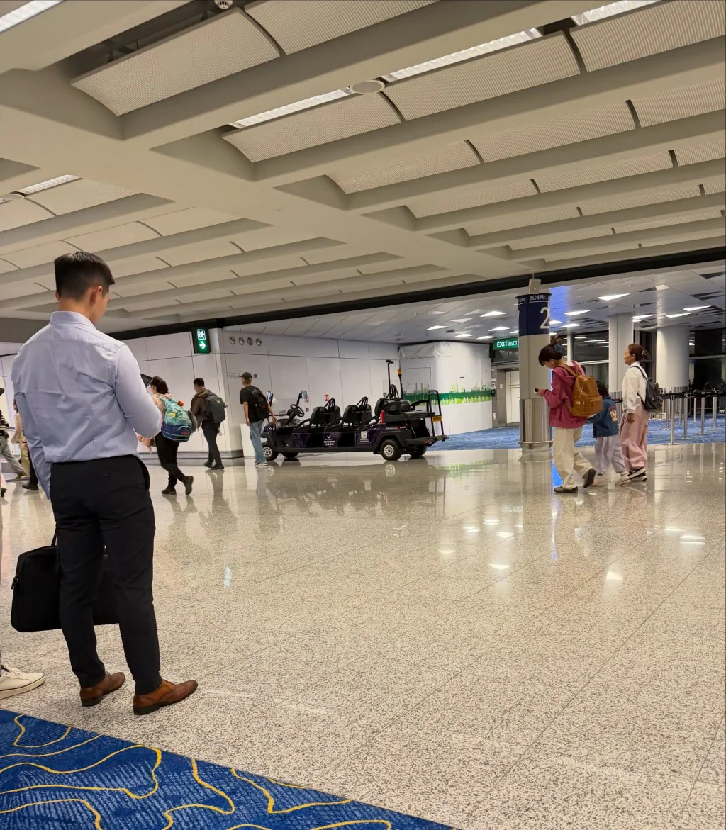 鄧龍威的兄長Billy在香港國際機場大堂等候接機，現場有多名途人及一輛機場電動車經過。