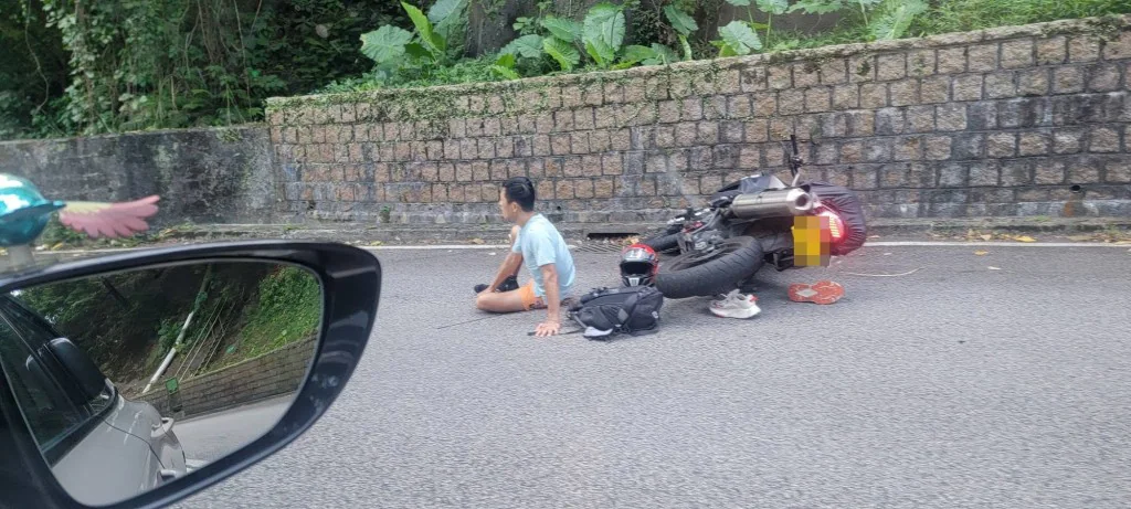 一名男子坐在荃錦公路路邊，身旁有倒地的電單車及頭盔，背景為石牆及樹木。
