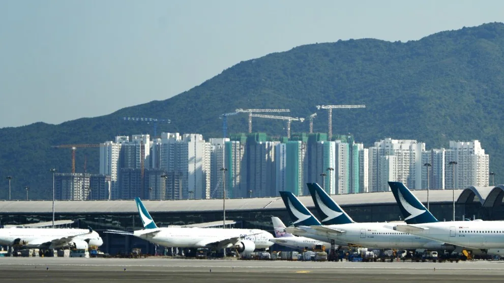 香港國際機場停機坪上停泊的國泰航空客機，背景可見遠處的住宅大廈群及山巒。