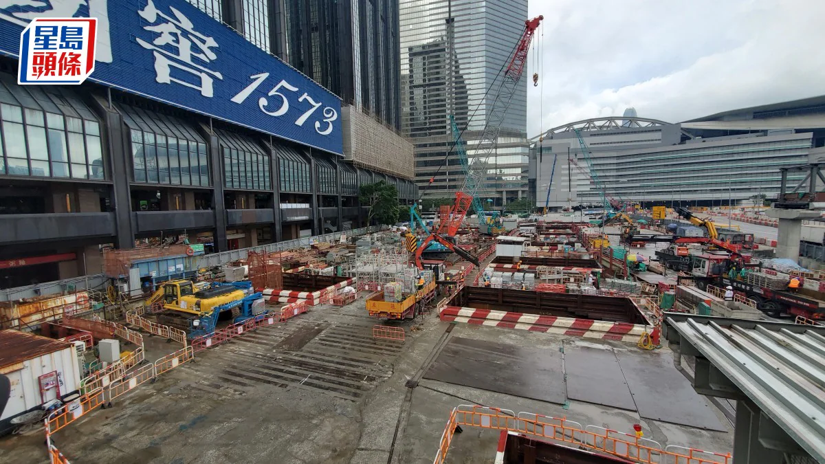 灣仔大型建築工地現場，可見多部起重機、挖掘機及工程車輛，背景為香港會議展覽中心及周邊商業大廈。